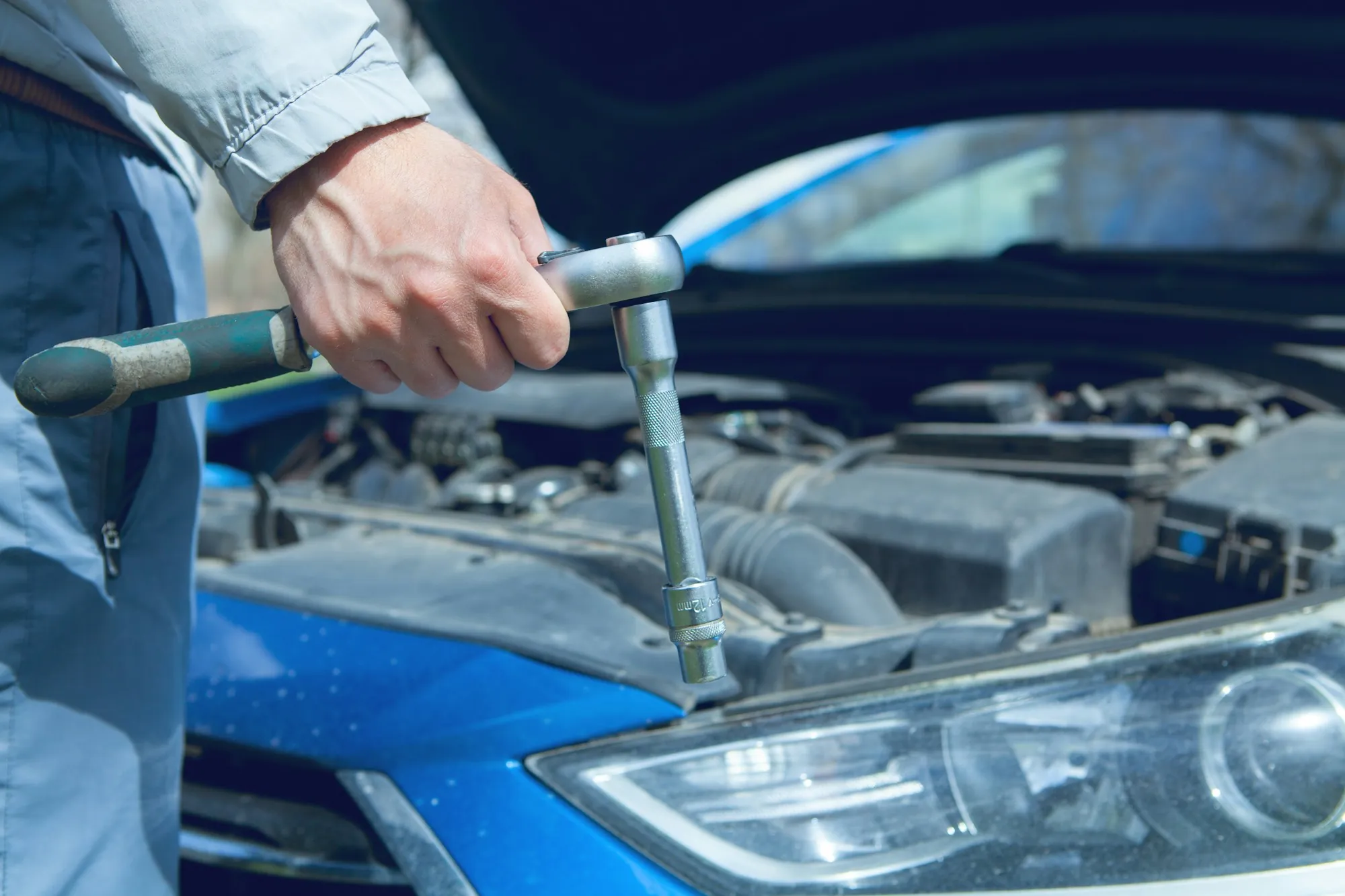 Hand turning a ratchet on an engine beside a blue car fender